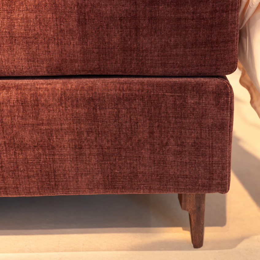 Close-up of a bed frame upholstered in textured rust-brown fabric, with white bedding and a visible wooden leg against a light neutral floor.