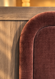 Close-up of a bed headboard with a warm wood panel on the left and a rounded upholstered section on the right in textured rust-brown fabric, lit by soft warm sunlight.