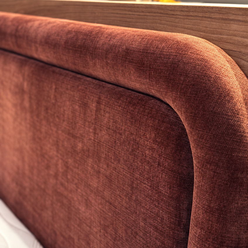 Close-up of a upholstered bed headboard in a warm rust-brown fabric, showing the curved top rail and textured fabric detail against a white mattress and wood headboard frame.